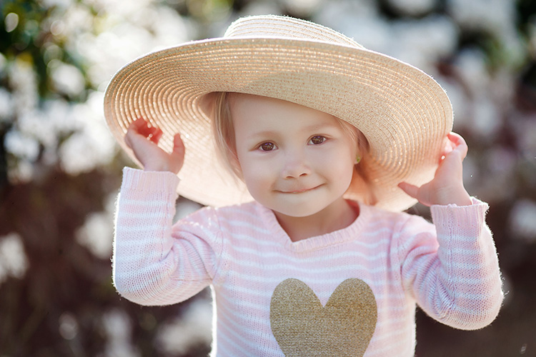 Ein junges Kind trägt einen breiten Strohhut und lächelt in die Kamera. Es trägt einen rosa Pullover mit einem großen, herzförmigen Muster auf der Brust. Das Kind befindet sich draußen an einem sonnigen Tag, umgeben von unscharfem Hintergrund aus hellem Grün. Der Strohhut bietet Schutz vor der Sonne, was auf den Kontext von Arbeitsschutz und Prävention durch angemessene Bekleidung und Sonnenschutz hinweist, relevant für die Unfallkasse NRW.