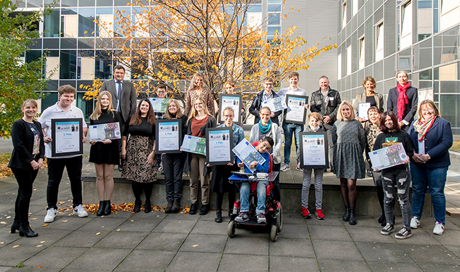 Das Bild zeigt eine Gruppe von 21 Personen, die vor einem modernen Gebäude mit großen Fenstern posiert. Einige Personen halten gerahmte Auszeichnungen. Im Hintergrund ist ein Baum mit Herbstlaub zu sehen. Die Personen sind unterschiedlich gekleidet und es gibt eine Mischung aus Männern und Frauen. Im Vordergrund sitzt ein Kind in einem Rollstuhl. Die Veranstaltung hängt vermutlich mit Arbeitsschutz und Prävention im Kontext der Unfallkasse NRW zusammen.