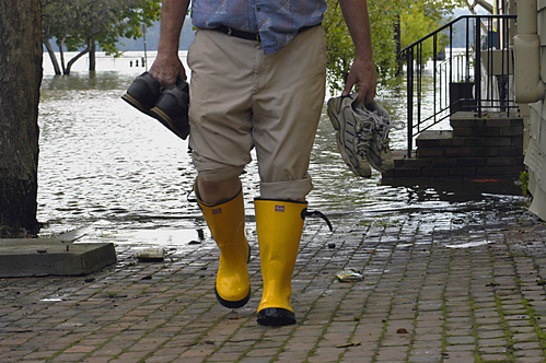 Eine Person in gelben Gummistiefeln geht durch eine überschwemmte Straße. In der linken Hand trägt sie ein Paar Schuhe und in der rechten Hand ein Paar Stiefel. Die Umgebung zeigt überflutetes Wasser auf einer Straße mit Bäumen im Hintergrund, die teilweise im Wasser stehen. Eine Treppe führt zu einem Gebäude am rechten Bildrand. Der Kontext des Bildes bezieht sich auf Arbeitsschutz und Prävention bei Hochwassersituationen.