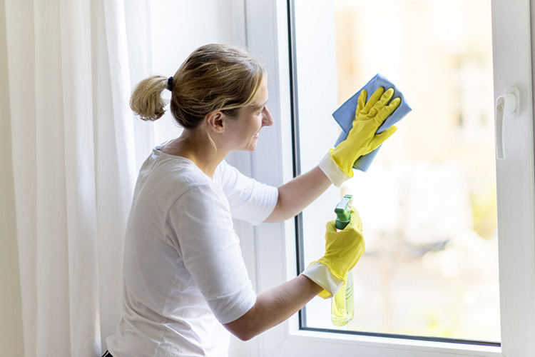 Eine Frau mit blonden Haaren, die zu einem Zopf gebunden sind, trägt gelbe Schutzhandschuhe und reinigt ein Fenster. Sie benutzt ein blaues Tuch in ihrer rechten Hand und hält einen grünen Sprühreiniger in ihrer linken Hand. Die Frau steht vor einem hellen Hintergrund und trägt ein weißes Oberteil. Das Bild vermittelt den Kontext von Arbeitsschutz und Prävention im Rahmen der Unfallkasse NRW, indem auf sichere und ergonomische Arbeitspraktiken bei der Fensterreinigung hingewiesen wird.