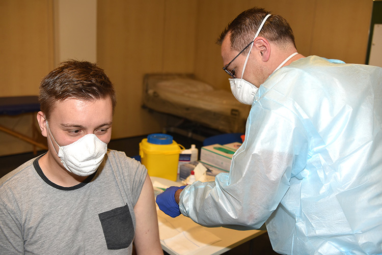 Ein Mann sitzt und wird von einem Angehörigen des medizinischen Personals geimpft. Beide tragen OP-Masken. Im Hintergrund sind medizinische Materialien und eine Liege zu sehen. Dieses Bild könnte im Kontext von Arbeitsschutz und Präventionsmaßnahmen verwendet werden, um die Wichtigkeit von Impfungen als Teil der Gesundheitsprävention hervorzuheben.