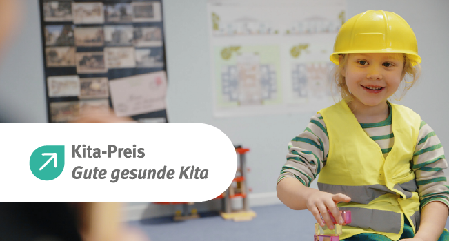 Ein kleines Kind sitzt in einer Kita-Umgebung und trägt einen gelben Bauhelm sowie eine gelbe Warnweste über einem gestreiften Pullover. Im Hintergrund sind spielerische Baupläne an der Wand zu sehen. Links im Bild befindet sich ein Logo mit der Aufschrift 'Kita-Preis Gute gesunde Kita'. Das Bild vermittelt Informationen zum Thema Arbeitsschutz und Prävention in Kindertagesstätten.