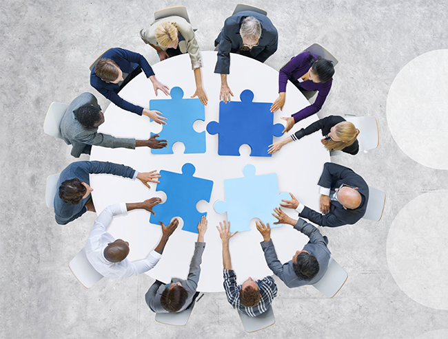 Mehrere Personen unterschiedlicher Herkunft sitzen an einem runden Tisch und fügen große, blaue Puzzleteile zusammen. Die Szene symbolisiert Teamarbeit und Zusammenarbeit, wichtige Aspekte im Bereich des Arbeitsschutzes und der Prävention, wie sie von der Unfallkasse NRW gefördert werden. Der Hintergrund ist neutral gehalten, damit der Fokus auf den Personen und den Puzzleteilen liegt.