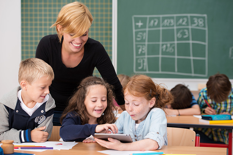 Eine Lehrerin mit kurzen blonden Haaren steht lächelnd hinter drei Grundschulkinder, die gemeinsam an einem Tisch sitzen. Zwei Mädchen und ein Junge betrachten interessiert ein Tablet, das ein Mädchen in der Hand hält. Im Hintergrund ist eine grüne Tafel mit Kreidezeichnungen, darauf schemenhaft angedeutet ein Zahlenraster, zu erkennen. Weitere Kinder sitzen weiter hinten an Tischen und scheinen ebenfalls beschäftigt zu sein. Das Bild vermittelt eine Atmosphäre von interaktivem und digital unterstütztem Lernen in einer Grundschule und kann im Kontext von Arbeitsschutz und Prävention auf die Förderung von Bildung und Sicherheit im Schulalltag hinweisen.