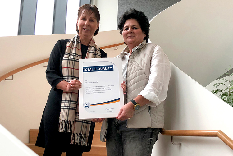 Zwei Frauen stehen auf einer modernen Treppe mit Holzgeländer und halten gemeinsam eine Urkunde mit der Aufschrift 'Total E-Quality'. Die Urkunde könnte für Gleichstellung oder Diversität im Arbeitsumfeld stehen. Die Umgebung wirkt professionell und hell, was auf ein Bürogebäude hinweist. Dies könnte mit dem Thema Arbeitsschutz und Prävention im Zusammenhang mit der Unfallkasse NRW stehen.