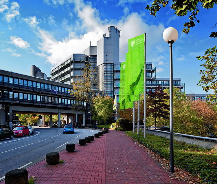 Das Bild zeigt das Hauptgebäude der Unfallkasse NRW an einem sonnigen Tag. Es befindet sich in einer städtischen Umgebung mit modernen architektonischen Elementen. Vor dem Gebäude steht ein großes, grünes Banner mit dem Logo und Schriftzug der Unfallkasse NRW. Der Himmel ist teilweise bewölkt, und Herbstlaub ist auf den Bäumen zu sehen. Ein Gehweg mit roten Pflastersteinen führt zum Gebäude, daneben sind eine Straße und geparkte Fahrzeuge zu sehen.