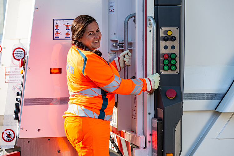 Eine Frau in leuchtend orangefarbener Warnkleidung und weißen Handschuhen bedient die Hebevorrichtung eines Müllfahrzeugs. Auf der Fahrzeugseite sind farbige Knöpfe und Sicherheitshinweise angebracht. Die Szene betont die Bedeutung von Arbeitsschutz und Prävention im Berufsalltag, insbesondere im Straßenverkehr und bei der Müllentsorgung. Das Bild könnte für die Unfallkasse NRW relevant sein, da es die Einhaltung von Sicherheitsvorschriften und das Tragen von Warnkleidung verdeutlicht.