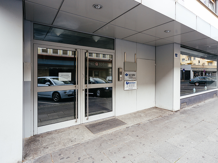 Der Eingangsbereich eines modernen Bürogebäudes mit zwei großen Glastüren. Neben den Türen ist ein Schild der Unfallkasse NRW sichtbar. Die Fassade besteht aus metallischen Paneelen. Eine Fußmatte liegt vor den Türen. Auf der gegenüberliegenden Straßenseite sind Autos und Geschäfte zu sehen. Dies könnte auf einen städtischen Standort hinweisen.
