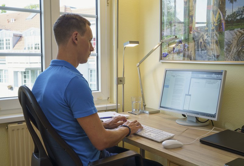 Ein Mann sitzt auf einem ergonomischen Bürostuhl an einem Schreibtisch. Vor ihm steht ein großer Monitor, auf dem Text bearbeitet wird. Neben dem Monitor stehen eine Lampe und ein Glas Wasser. Im Hintergrund ist ein Fenster mit Blick auf andere Gebäude sowie ein buntes Poster an der Wand sichtbar. Das Bild thematisiert Arbeitsschutz und Prävention, indem es auf die ergonomische Gestaltung des Arbeitsplatzes hinweist.