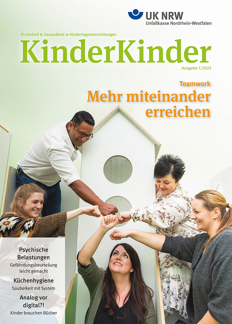 Eine Gruppe von vier Erwachsenen, zwei Männer und zwei Frauen, interagiert positiv und hebt gemeinsam ihre Hände in die Luft. Sie stehen um ein weißes, hausähnliches Kulissenelement mit runden Fenstern. Der Hintergrund ist farblich dezent gehalten. Der Fokus liegt auf Teamwork und Zusammenarbeit in der Arbeitswelt. Oben sind die Logos der Unfallkasse Nordrhein-Westfalen und die Überschrift der Zeitschrift 'KinderKinder' zu sehen. Zusätzliche Themen auf dem Cover sind psychische Belastungen, Küchenhygiene und Digitalisierung in Kindertagesstätten.