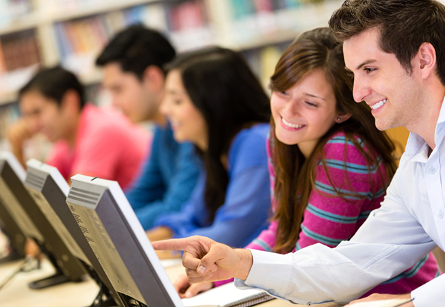 Fünf junge Erwachsene sitzen in einer Reihe an Computern in einer Bibliothek. Drei Männer und zwei Frauen arbeiten konzentriert. Zwei der Personen lächeln und einer zeigt auf einen Bildschirm. Im Hintergrund sind Bücherregale zu sehen. Diese Szene könnte auf die Bedeutung von Bildung und Verantwortung im Rahmen des Arbeitsschutzes hinweisen.