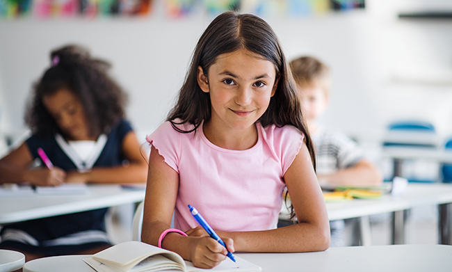 Ein junges Mädchen im Vordergrund sitzt an einem Tisch in einem Klassenzimmer und schreibt in ein Heft. Sie trägt ein rosa T-Shirt und hält einen blauen Stift in der Hand. Im Hintergrund sind zwei weitere Kinder zu sehen, ein Junge und ein Mädchen, die ebenfalls an Tischen sitzen und schreiben. Die Umgebung ist hell und ordentlich, passend zum Thema Bildung und Lernen.