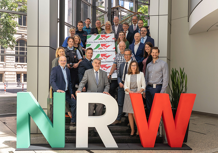 Eine Gruppe von Personen steht auf einer Treppe hinter großen, dekorativen Buchstaben 'NRW', die in den Farben Grün, Weiß und Rot gehalten sind. Einige Teilnehmerinnen und Teilnehmer halten ein Banner mit der Aufschrift 'SicherImDienst', das auf Arbeitsschutz hinweist. Die Szene spielt in einem modernen Gebäude mit Glaswänden, das viel Tageslicht einlässt. Die Gruppe scheint aus verschiedenen Berufsgruppen zu bestehen, darunter auch eine Person in Polizeiuniform. Diese Versammlung steht im Kontext von Unfallprävention und Arbeitsschutz, einem wichtigen Thema der Unfallkasse NRW.