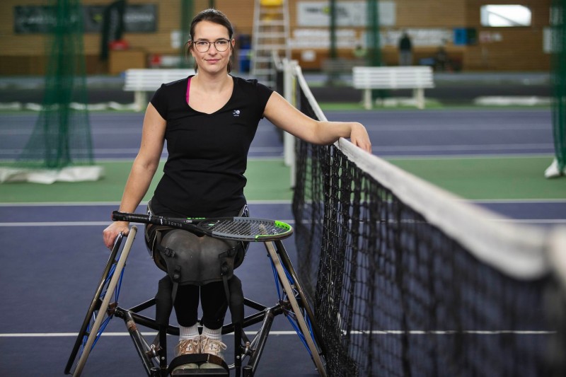 Eine Frau im Rollstuhl lächelt und stützt sich auf ein Tennisnetz in einer Sporthalle. Sie trägt sportliche Kleidung und hat einen Schläger auf ihrem Schoß. Der Hintergrund zeigt einen Tennisplatz mit weiteren Plätzen, Netz und Sportausrüstung. Diese Szene betont Inklusion und Barrierefreiheit im Sport. Kontext: Arbeitsschutz und Prävention sind auch im Behindertensport wichtig, bei dem das Risiko von Verletzungen minimiert werden muss.