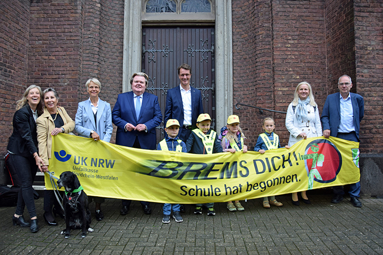 Eine Gruppe von acht Erwachsenen und vier Kindern steht vor einem Backsteingebäude. Sie halten ein gelbes Banner mit der Aufschrift 'BREMS DICH! Schule hat begonnen.' Kinder tragen gelbe Kappen. Auf dem Banner befindet sich das Logo der Unfallkasse NRW und ein Bild eines bremsenden Autos. Links ist ein Blindenführhund zu sehen. Die Szene deutet auf eine Präventionsaktion zum Schulbeginn hin.
