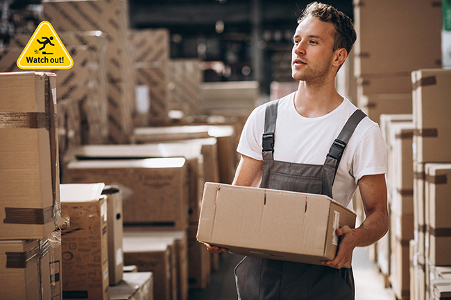 Ein Mann in Arbeitskleidung trägt einen Karton durch eine Lagerhalle mit vielen gestapelten Kartons. Oben links ist ein gelbes Warnschild mit der Aufschrift 'Watch out!' und einem Symbol für Rutschgefahr sichtbar. Dies deutet auf das Thema Arbeitssicherheit und Prävention von Unfällen hin, im Kontext der Unfallkasse NRW.