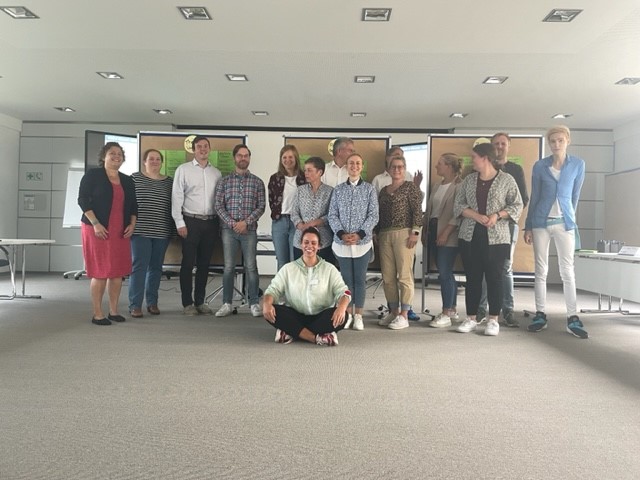 Ein Dutzend Menschen stehen und eine Person sitzt in einem modernen Konferenzraum mit hellen Wänden und Teppichboden. Im Hintergrund sind zwei Pinnwände sichtbar. Die Gruppe lächelt und wirkt entspannt, einige Personen sind leger gekleidet. Diese Art der Versammlung könnte im Kontext von Arbeitsschutz und Prävention der Unfallkasse NRW stehen, bei der Themen wie Sicherheit am Arbeitsplatz oder Gesundheitsförderung diskutiert werden könnten.