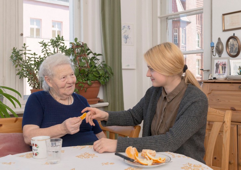 In einem hellen Raum mit Pflanzen und Bildern im Hintergrund sitzen eine ältere Frau und eine jüngere Frau an einem Tisch. Die jüngere Frau in grauer Strickjacke reicht der älteren Frau ein Stück Orange. Auf dem Tisch liegt ein Teller mit weiteren Orangenscheiben und ein Glas Wasser. Die Szene vermittelt eine freundliche und fürsorgliche Interaktion, die den sozialen Aspekt von Prävention und Gemeinschaftsunterstützung im Zusammenhang mit Arbeitsschutz und der Unfallkasse NRW betont.
