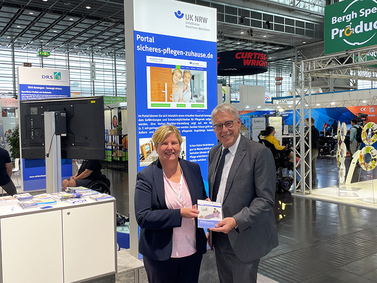 Zwei Personen stehen an einem Messestand der Unfallkasse NRW. Im Hintergrund ist ein großer Bildschirm mit Informationen über das Portal 'sicheres-pflegen-zuhause.de' zu sehen. Der Stand ist Teil einer Messehalle mit verschiedenen Ausstellern, die sich auf Arbeitsschutz und Prävention konzentrieren. Broschüren und Informationsmaterial liegen auf einem Tisch bereit.
