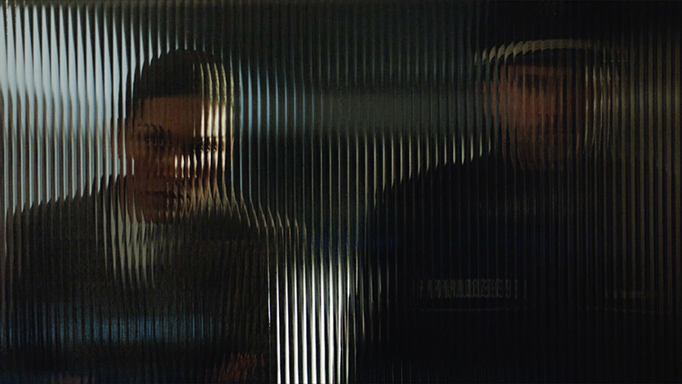 Zwei Personen stehen hinter einer geriffelten Glasscheibe und sind unscharf zu erkennen. Links eine Person in heller Kleidung, rechts eine in dunkler Uniform. Die Unschärfe symbolisiert möglicherweise Barrieren oder Distanz, die auch im Kontext von Arbeitsschutz und Prävention der Unfallkasse NRW relevant sind.