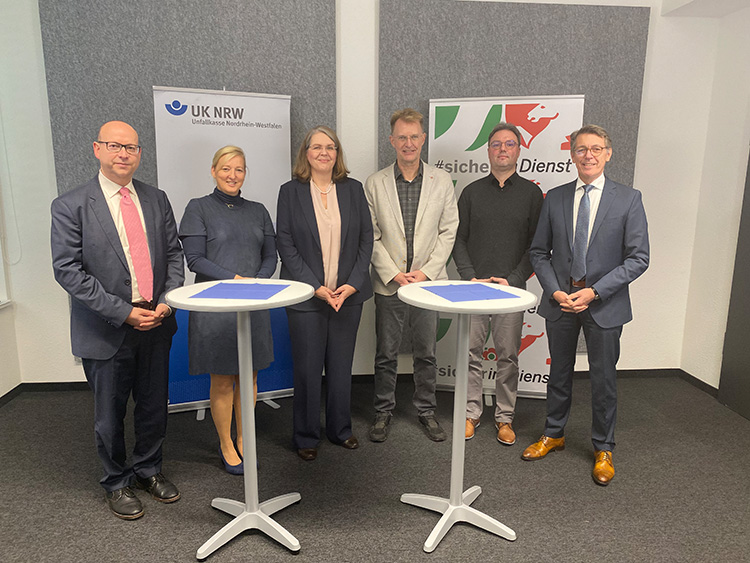 Sechs Personen stehen in formeller Kleidung in einem Innenraum vor zwei Bannern der Unfallkasse Nordrhein-Westfalen. Sie stehen hinter zwei weißen Stehtischen, auf denen blaue Mappen liegen. Die Szene deutet auf eine formelle Veranstaltung oder eine Pressekonferenz hin, die sich mit Arbeitsschutz und Prävention beschäftigt.