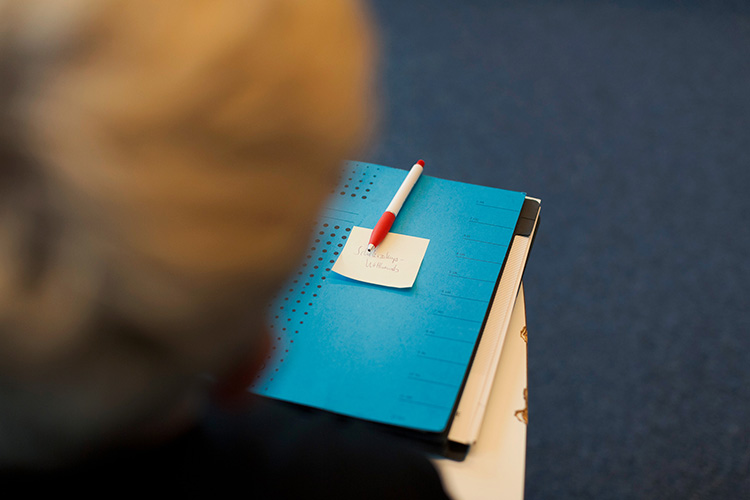 Eine Person in einem formellen Umfeld hält ein blaues Notizbuch auf dem ein roter und weißer Kugelschreiber liegt. Darauf ist ein Zettel mit handschriftlicher Notiz befestigt. Der Fokus liegt auf der Organisation und Vorbereitung, möglicherweise im Kontext einer Schulung oder Besprechung zur Förderung von Arbeitsschutz und Prävention. Der Hintergrund ist unscharf, wodurch der Notizblock im Vordergrund hervorgehoben wird.