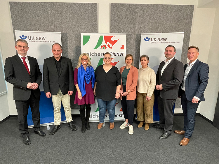 Acht Personen stehen in einem Büro vor Werbebannern der Unfallkasse Nordrhein-Westfalen. Zwei Banner tragen das Logo der UK NRW und vermitteln den Kontext von Arbeitsschutz und Prävention. Die Gruppe besteht aus vier Männern und vier Frauen, die entweder formelle oder semiförmelle Kleidung tragen. Der Hintergrund ist schlicht, mit grauen und weißen Wänden.