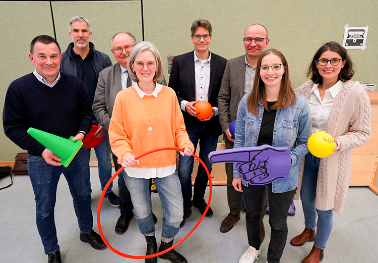 Acht Personen stehen in einer Sporthalle und halten bunte Trainingsmaterialien wie Kegel, Reifen und Bälle. Die Gruppe besteht aus Männern und Frauen mittleren Alters, die in lockerer Freizeitkleidung gekleidet sind. Sie lächeln und blicken in die Kamera. Im Hintergrund sind Sprossen- und Weichbodenmatten zu sehen. Das Szenario unterstützt das Thema Arbeitsschutz und Prävention im Kontext von sportlichen Aktivitäten.