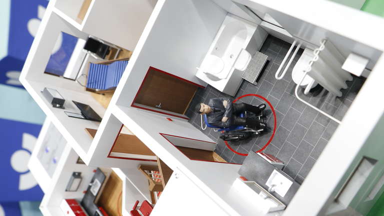 Das Bild zeigt ein Modell eines barrierefreien Badezimmers mit einem Rollstuhlfahrer. Im Raum befinden sich Haltegriffe neben der Toilette und eine bodenebene, offene Dusche mit Duschvorhang. Der Raum ist geräumig, um Bewegung im Rollstuhl zu ermöglichen. Diese Darstellung dient als Beispiel für barrierefreies Design im Kontext von Arbeitsschutz und Prävention, gefördert von der Unfallkasse NRW.