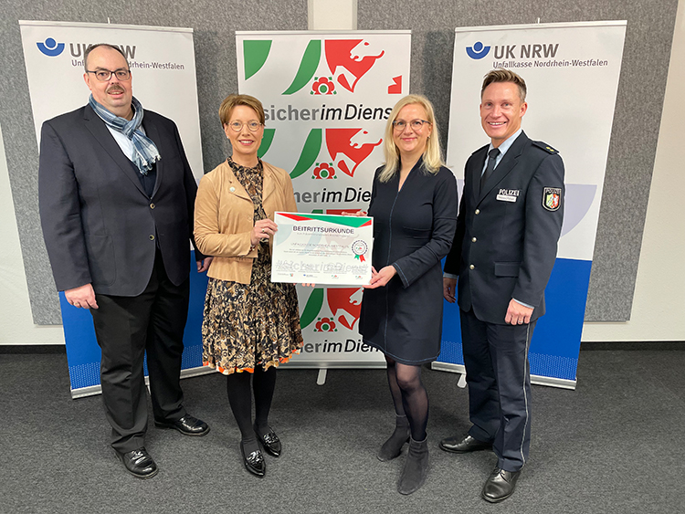 Vier Personen stehen nebeneinander und halten eine Sicherheitsurkunde. Zwei der Personen tragen Anzüge, eine Person trägt eine Uniform und eine Person ein Kleid mit einem Blazer. Im Hintergrund sind zwei Roll-ups mit den Logos der Unfallkasse Nordrhein-Westfalen und dem Slogan 'Sicher im Dienst' zu sehen. Die Szene vermittelt den Kontext von Arbeitsschutz und Prävention, wie es für die Unfallkasse NRW typisch ist.