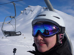 Eine Person mit einem weißen Skihelm und einer reflektierenden Skibrille sitzt in einem Sessellift. Im Hintergrund sind schneebedeckte Berge unter einem klaren blauen Himmel zu sehen. Dieses Bild unterstreicht die Bedeutung von Sicherheitsausrüstung beim Skifahren, ein wichtiges Thema für die Unfallkasse NRW im Bereich Arbeitsschutz und Prävention.
