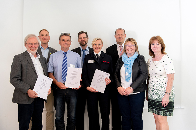 Acht Personen stehen nebeneinander vor einem weißen Hintergrund. Drei Personen in der Mitte halten Urkunden in den Händen. Die Gruppe besteht aus vier Männern und vier Frauen, die formell gekleidet sind. Der Kontext deutet auf eine Veranstaltung der Unfallkasse NRW hin, die sich auf Arbeitsschutz und Prävention konzentriert.