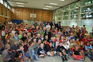 Eine große Gruppe von Kindern versammelt sich in einer Turnhalle mit Fenstern und Backsteinwänden. Einige Erwachsene stehen im Hintergrund. Der Raum wirkt lebhaft und fröhlich. Diese Szene könnte in Zusammenhang mit einem Schul- oder Freizeitprogramm stehen, das das Bewusstsein für Arbeitsschutz und Prävention im Alltag fördert, in Zusammenarbeit mit der Unfallkasse NRW.