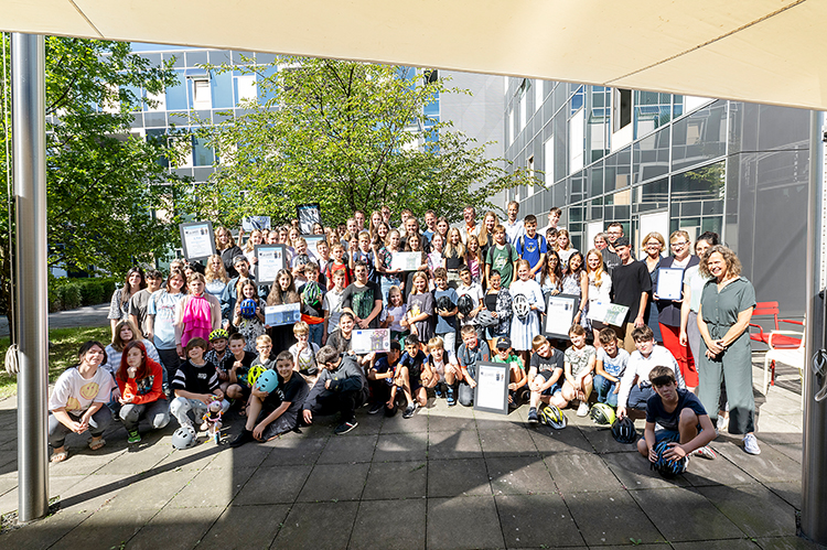 Eine große Gruppe von Kindern und einigen Erwachsenen posiert mit Urkunden. Sie stehen draußen auf einem gepflasterten Bereich vor einem modernen Gebäude. Das Bild könnte im Rahmen einer Veranstaltung zur Arbeitssicherheit oder Prävention aufgenommen worden sein. Die Umgebung ist von Bäumen umgeben, und es scheint ein sonniger Tag zu sein. Die Anwesenheit der Erwachsenen deutet auf eine offizielle oder pädagogische Veranstaltung hin.