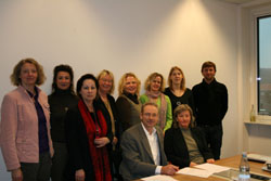 Eine Gruppe von zehn Personen, sieben Frauen und drei Männer, posieren in einem Büro. Zwei Personen sitzen an einem Tisch im Vordergrund, während die anderen dahinter stehen. Auf dem Tisch liegen Papiere und eine Wasserflasche ist sichtbar. Der Raum hat weiße Wände und ein Fenster rechts. Das Bild vermittelt ein formelles Meeting, möglicherweise im Kontext von Arbeitsschutz oder Prävention für die Unfallkasse NRW.