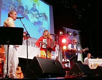 Vier Musiker performen auf einer Bühne. Eine Frau spielt Akkordeon und singt, ein Mann spielt Gitarre und singt, ein weiterer spielt Bass und ein Mann sitzt am Keyboard. Im Hintergrund ist eine Projektion mit Kindern auf der Leinwand sichtbar. Die Szene scheint Teil einer Veranstaltung zur Förderung von Sicherheit und Prävention zu sein, möglicherweise im Kontext der Unfallkasse NRW.