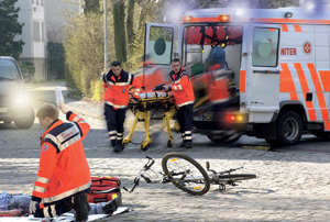 Zwei Rettungssanitäter in leuchtend oranger Kleidung schieben eine Trage in einen geparkten Rettungswagen mit geöffneten Türen. Vor ihnen liegt ein Fahrrad umgestürzt auf dem Kopfsteinpflaster. Ein weiterer Sanitäter kniet neben einem Verletzten, der auf dem Boden liegt. Im Hintergrund sind Bäume und Gebäude sichtbar. Die Szene steht im Kontext des Arbeitsschutzes und der Unfallprävention, indem sie die schnelle Reaktion bei einem Unfall darstellt.