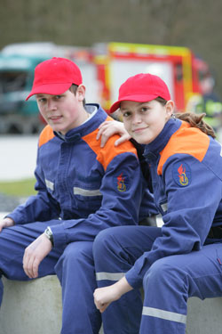 Zwei Jugendliche tragen blaue Uniformen mit orangefarbenen Schulterpartien und rote Kappen. Sie sitzen nebeneinander auf einer Betonmauer und wirken entspannt. Im Hintergrund sind Fahrzeuge unscharf zu erkennen, darunter ein Feuerwehrauto. Das Bild könnte in Zusammenhang mit einem Arbeitsschutz- oder Präventionstraining für Jugendgruppen der Unfallkasse NRW stehen.