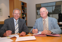 Zwei ältere Männer sitzen an einem Holztisch in einem Büro. Beide tragen Anzüge; der Mann links hat eine Brille auf und lächelt, während er mit einem Stift auf ein Dokument zeigt. Der Mann rechts hält einen blauen Stift über ein geöffnetes Aktenbuch. Im Hintergrund sind Regale mit Büchern und ein großes Fenster, das Tageslicht hereinlässt. Das Bild könnte im Kontext von Arbeitsschutz und Prävention bei der Unfallkasse NRW stehen, da es eine professionelle Arbeitsumgebung zeigt.