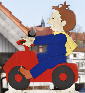 Eine bemalte Holzfigur eines Kindes in blauer Kleidung mit gelbem Schal sitzt auf einem roten Spielzeugtraktor. Die Figur befindet sich vor einem Hintergrund mit Gebäuden, die an eine ländliche Umgebung erinnern. Diese Darstellung symbolisiert Arbeitssicherheit und Prävention im Kinderspielbereich im Kontext der Unfallkasse NRW.