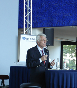 Ein Mann im Anzug hält ein Mikrofon und spricht bei einer Veranstaltung der Unfallkasse NRW. Im Hintergrund steht ein Banner mit dem Logo der Unfallkasse NRW. Neben ihm auf dem Tisch stehen zwei Wasserflaschen, und im Raum sind große Fenster sichtbar, die natürliches Licht hereinlassen. Diese Veranstaltung könnte sich auf Themen wie Arbeitsschutz und Prävention konzentrieren.