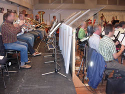 Das Bild zeigt eine Orchesterprobe mit Musikern, die Blas- und Streichinstrumente spielen. Plexiglas-Trennwände zwischen den Reihen dienen als Schutzmaßnahme zur Lärmreduzierung und hygienischen Prävention. Die Musiker sitzen in einem Probenraum auf schwarzen Stühlen und verwenden Notenständer. Die Trennwände sind transparent und sorgsam zwischen den Musikern platziert. Diese Maßnahme kann im Kontext des Arbeitsschutzes und der Prävention bei der Unfallkasse NRW relevant sein, um die Sicherheit und Gesundheit der Mitarbeiter zu gewährleisten.