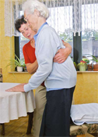 Eine Pflegekraft in einem roten Oberteil hilft einem älteren Mann in einem blauen Pullover beim Gehen. Sie unterstützt ihn mit einem Arm um seinen Rücken in einem häuslichen Umfeld. Im Hintergrund sind Zimmerpflanzen und Gardinen zu sehen. Das Bild thematisiert Arbeitsschutz und Prävention bei der Unterstützung von Senioren, um Unfälle im Pflegealltag zu vermeiden.