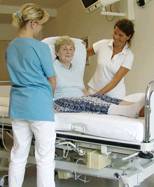 Zwei Pflegerinnen in medizinischer Kleidung assistieren einer älteren Dame beim Transfer in ein Krankenhausbett. Eine Pflegerin richtet die Kissen hinter der Frau, während die andere am Fußende des Bettes steht und Anweisungen gibt. Die Szene findet in einem gut beleuchteten Krankenhauszimmer statt und betont die Bedeutung von sicherem Patiententransfer, einem wichtigen Aspekt des Arbeitsschutzes und der Prävention im Gesundheitswesen.