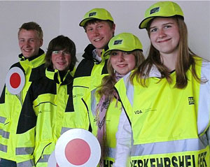 Das Bild zeigt fünf junge Menschen, die helle Warnwesten und Mützen tragen. Zwei von ihnen halten Verkehrskellen. Die Kleidung ist mit dem Aufdruck der Unfallkasse NRW versehen, was auf Arbeitsschutz und Präventionsmaßnahmen im Straßenverkehr hinweist. Die Gruppe lächelt und steht vor einem neutralen Hintergrund. Dies könnte eine Schulung oder ein Einsatz im Rahmen von Verkehrssicherheitsprogrammen darstellen, die von der Unfallkasse NRW unterstützt werden.