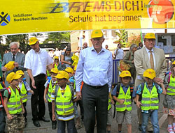 Eine Gruppe von Kindern trägt gelbe Warnwesten und Mützen. Sie werden von mehreren Erwachsenen begleitet. Alle stehen unter einem großen Banner mit der Aufschrift 'BREMS DICH! Schule hat begonnen', das von der Unfallkasse Nordrhein-Westfalen stammt. Die Szene spielt sich vor einem Schulgebäude ab, und die Aktion scheint auf Verkehrssicherheit und Prävention abzuzielen.