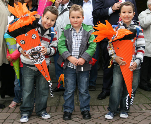 Drei Jungen stehen vor einer Gruppe von Erwachsenen, die teilweise im Bild sichtbar sind. Jeder der Jungen hält eine große, orangefarbene Schultüte in den Händen, die mit schwarzen und weißen Fußballmustern verziert ist. Die Jungen tragen lässige Kleidung mit Jeans und Jacken. Die Szene spielt wahrscheinlich am ersten Schultag, ein traditionelles Ereignis in Deutschland. Der Kontext könnte für die Unfallkasse NRW relevant sein im Sinne von Arbeitsschutz und Prävention, um auf die Sicherheit von Kindern auf Schulwegen hinzuweisen.