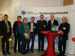 Eine Gruppe von Personen steht vor einem Infostand der Unfallkasse Nordrhein-Westfalen. Der Stand enthält Banner mit dem Logo und der Aufschrift 'Verkehrssicherheitsarbeit'. Auf einem Tisch ist ein Laptop platziert. Die Gruppe scheint in ein Gespräch vertieft zu sein und Broschüren sind sichtbar. Der Kontext deutet auf eine Veranstaltung zur Förderung von Sicherheit und Prävention im Verkehr hin.