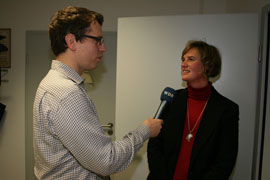 Ein Journalist mit Brille und kariertem Hemd hält ein Mikrofon mit einem Logo in der Hand und interviewt eine Frau. Die Frau trägt einen roten Rollkragenpullover und einen schwarzen Blazer. Sie stehen in einem Raum mit einer weißen Wand und einer teilweise geöffneten Tür im Hintergrund. Das Interview könnte sich um Themen wie Arbeitsschutz und Prävention bei der Unfallkasse NRW drehen.