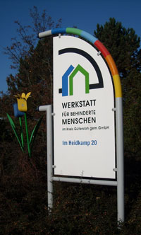 Ein großes weißes Schild steht vor einer Werkstatt für behinderte Menschen. Es trägt die Aufschrift 'Werkstatt für behinderte Menschen im Kreis Gütersloh gem. GmbH', sowie die Adresse 'Im Heidkamp 20'. Das Schild ist dekorativ mit einem geschwungenen bunten Element und einer stilisierten gelben Blume verziert. Der Hintergrund zeigt Bäume und einen klaren blauen Himmel. Der Kontext weist auf die Wichtigkeit von Inklusion und Arbeitsmöglichkeiten für Menschen mit Behinderungen hin, was für die Unfallkasse NRW relevant sein könnte im Sinne von Präventions- und Arbeitsschutzmaßnahmen.